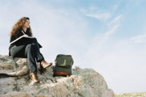 Jeune femme assise sur un rocher avec un sac à dos posé près d'elle et qui écrit dans un carnet.