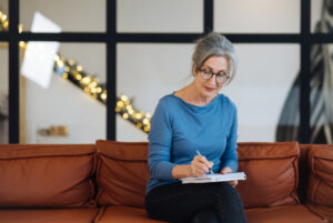 Femme âgée qui porte des lunettes, assise sur un canapé marron et qui écrit dans un cahier.