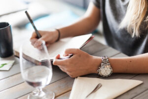 Femme qui écrit dans un cahier sur une table avec un verre posé à côté d'elle.