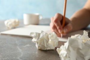 Personne qui écrit sur une feuille posée sur une table avec des boules de papier chiffonnées et une tasse de café.