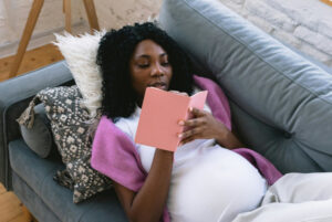 Femme enceinte allongée sur un canapé et qui écrit un dans un carnet rose.