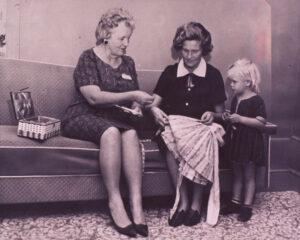 Photo ancienne représentant deux femmes âgées qui montrent à une petite fille comment coudre un vêtement.
