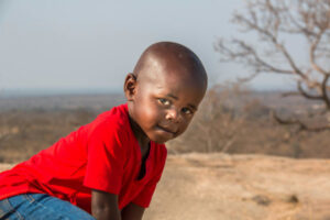 Enfant de couleur portant un tee-shirt rouge et regardant de face, à genoux au milieu de la savane.