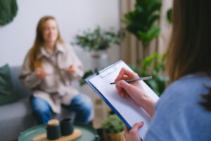 Jeune femme floutée assise face à une psychologue positionnée de dos et qui écrit dans un carnet.