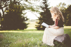 Jeune femme aux cheveux blonds longs, assise sur un banc dans un pré et qui lit un livre.