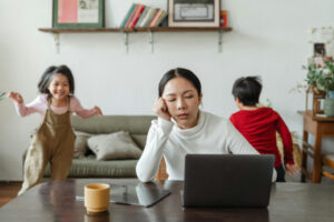 Deux enfants asiatiques qui courent dans le salon près de leur mère assise en face d'un ordinateur et qui essaie de se concentrer.
