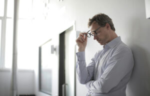 Homme qui porte une chemise, adossé à un mur, qui tient ses lunettes et est déprimé.