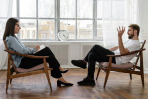 Homme assis sur une chaise et qui parle à une jeune femme également assise sur une chaise en face de lui, et qui prend des notes dans un cahier. 