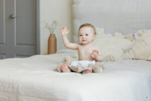 Bébé souriant vêtu d'une couche, assis sur un grand lit et qui lève la main.
