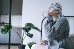 Femme atteinte d'un cancer du sein qui a un foulard sur les cheveux et boit un médicament assise sur son lit.